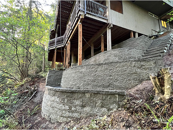 Multi-tiered block retaining wall with stairs on a steep slope behind a residential home.